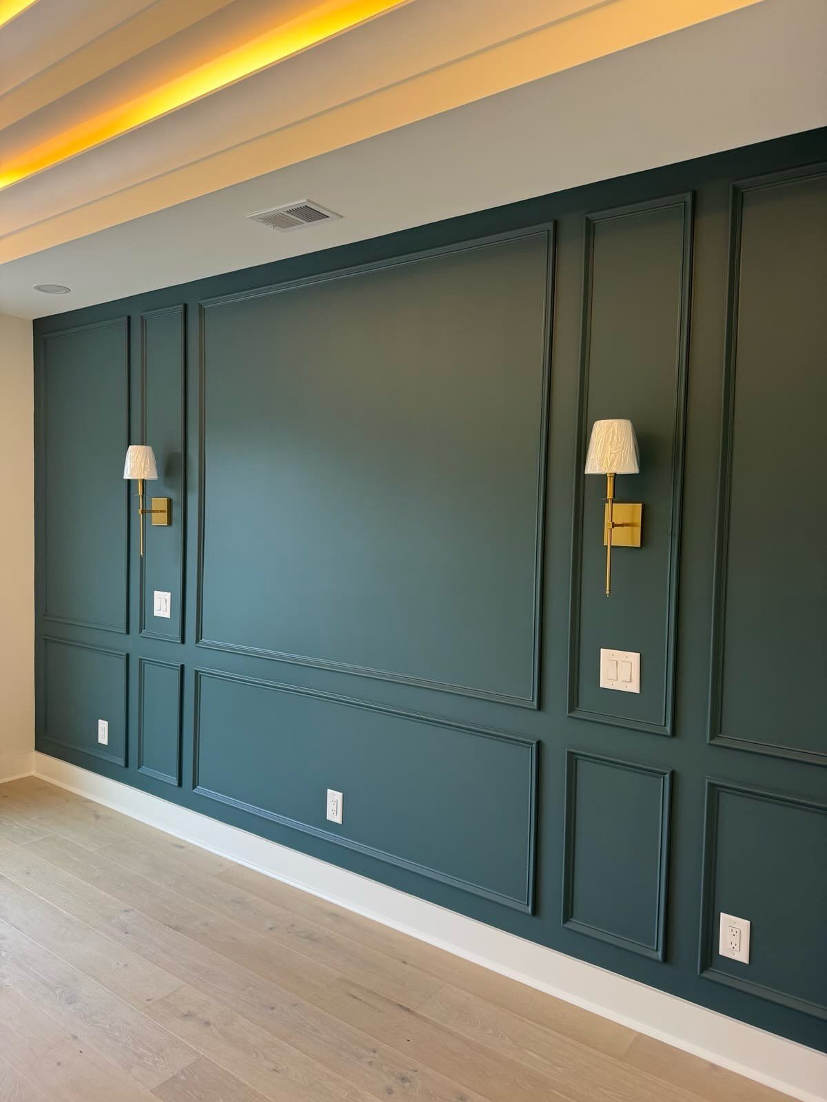 Dark teal wall with decorative picture frame molding and gold wall sconces above light wood floors.