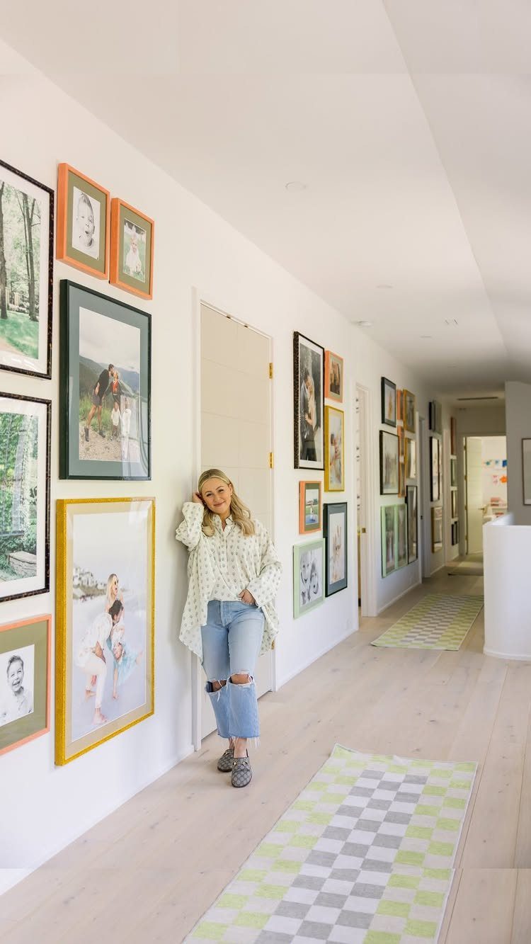 A woman leaning against a white wall decorated with a large, colorful gallery wall of framed photos.