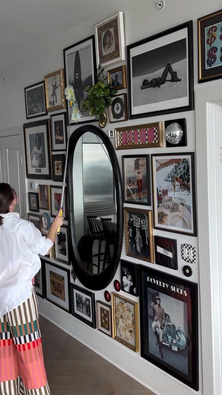 A person hanging a large oval mirror onto a wall filled with various framed art pieces.
