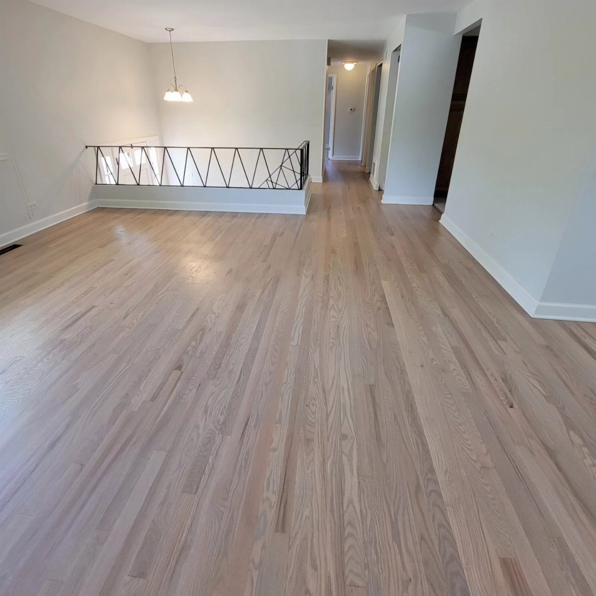 Light-colored hardwood flooring stretches across a large, empty room with white walls and a black railing.