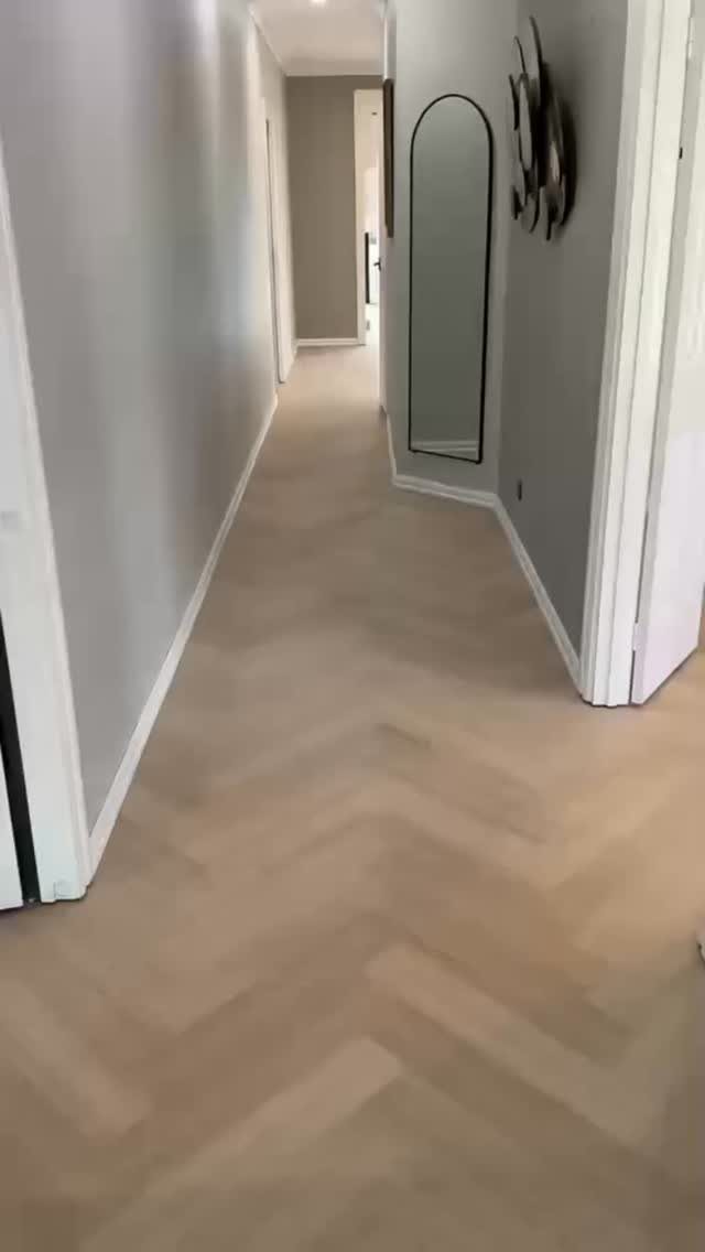 Light wood herringbone flooring running through a long, neutral-toned hallway with an arched mirror.