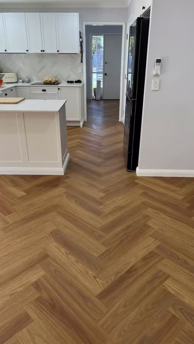 Herringbone patterned wooden flooring stretching through a modern white kitchen and into a hallway.