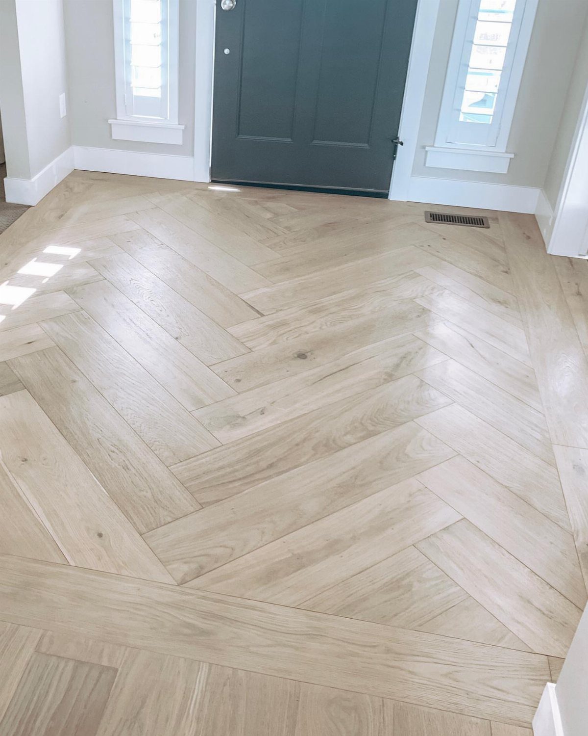 Light wood herringbone flooring leading towards a dark grey front door in a bright foyer.