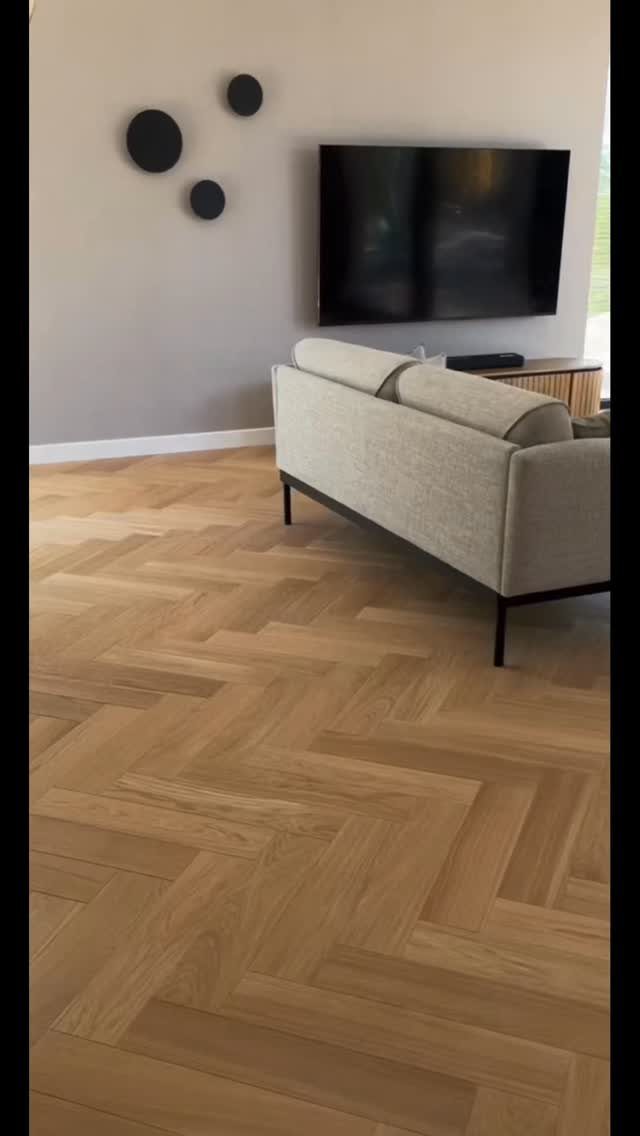 Light wood herringbone flooring in a modern living room with a beige sofa and wall-mounted television.