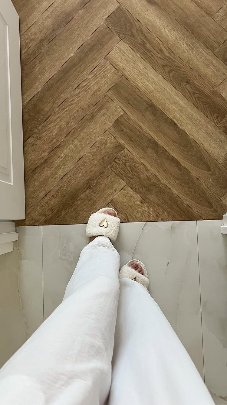 Person in white pants and slippers standing on white tile next to herringbone wood flooring.