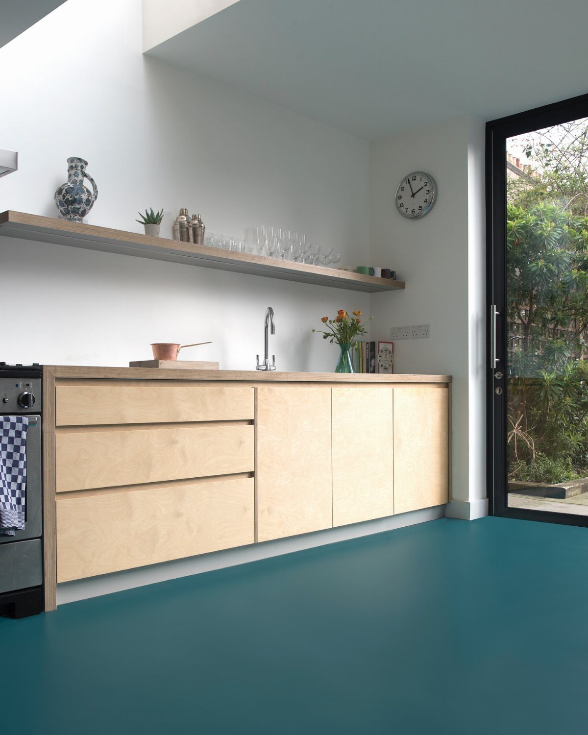 A modern kitchen with light wood cabinetry and a bold teal vinyl floor.