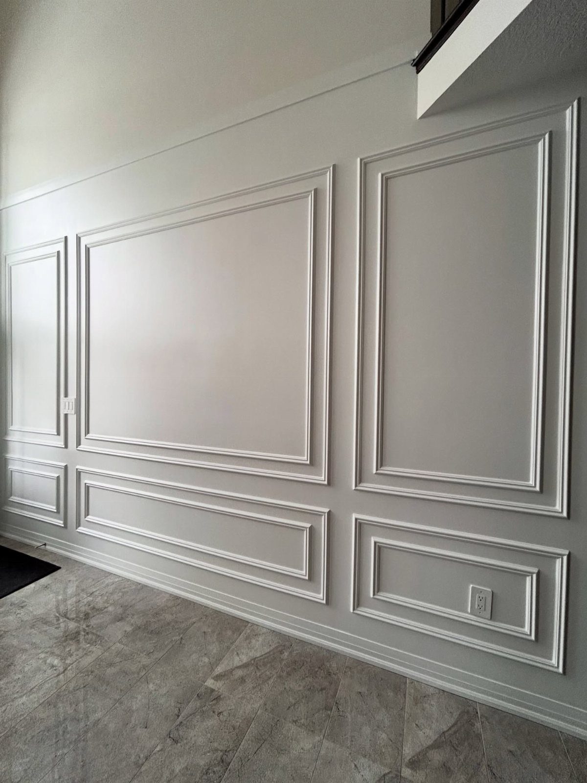 White decorative wall molding panels installed on a light gray entryway wall above stone flooring.