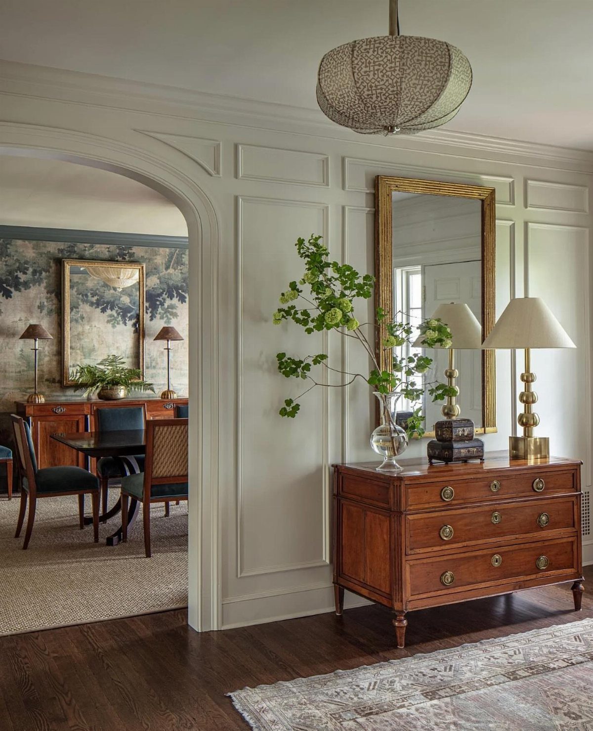 Light grey paneled wainscoting wall with a wooden dresser and gold mirror in an entryway.