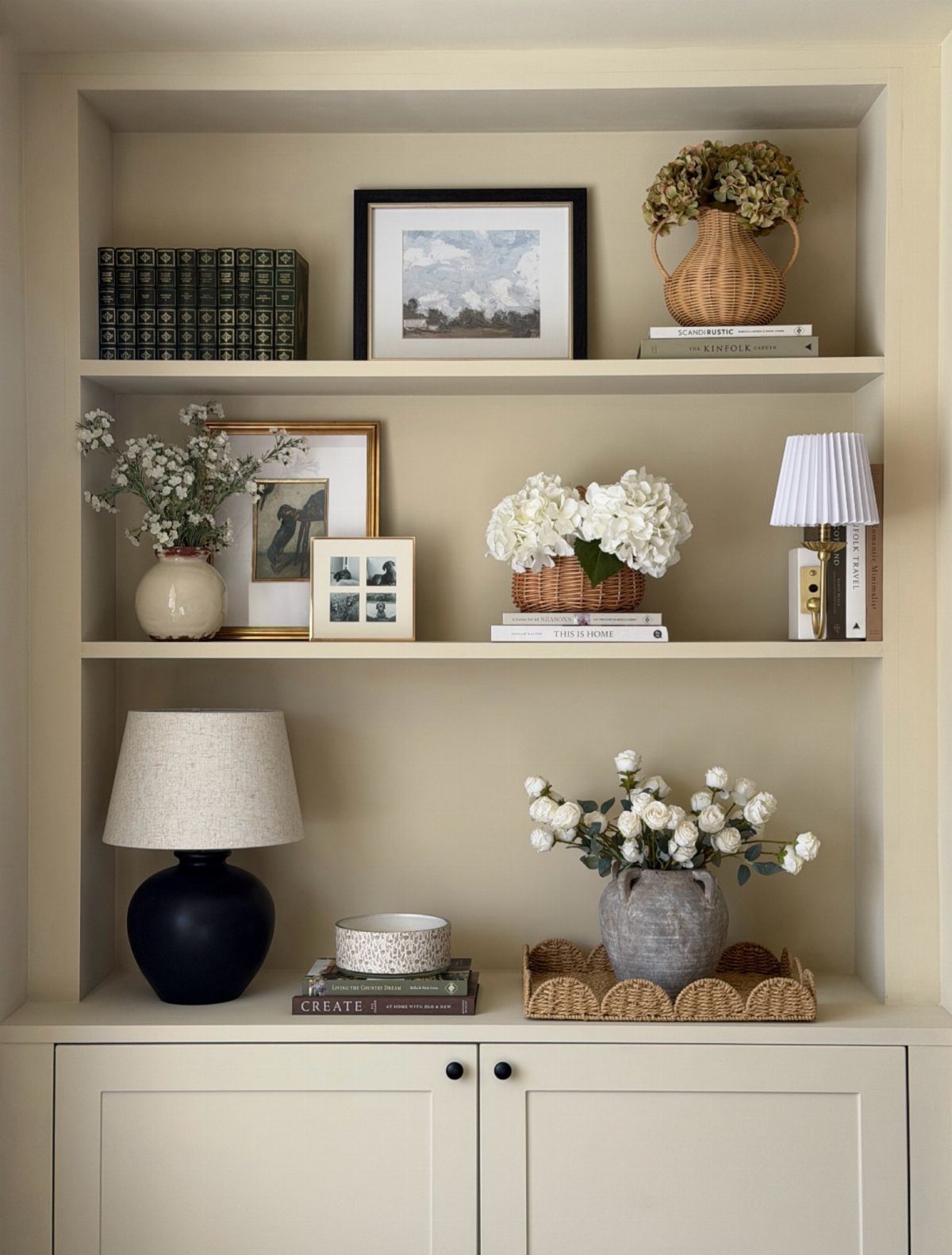 14 Wall Design in Living Room Ideas That Add Real Depth 14 Built-in beige shelves decorated with books, framed art, white flowers, and a small lamp.