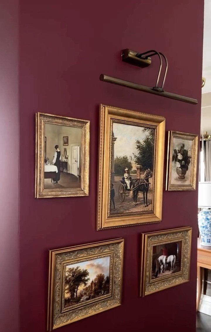 14 Wall Design in Living Room Ideas That Add Real Depth 10 Gold-framed paintings arranged on a deep burgundy wall with a brass picture light above them.
