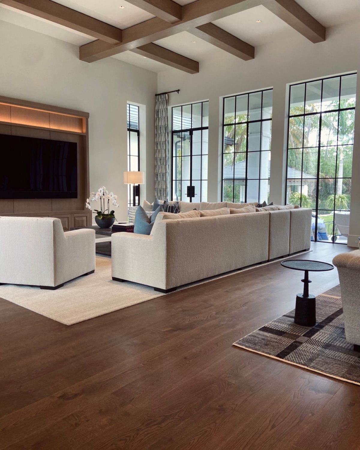 Mid-tone wood flooring in a modern living room with beige furniture and large windows.