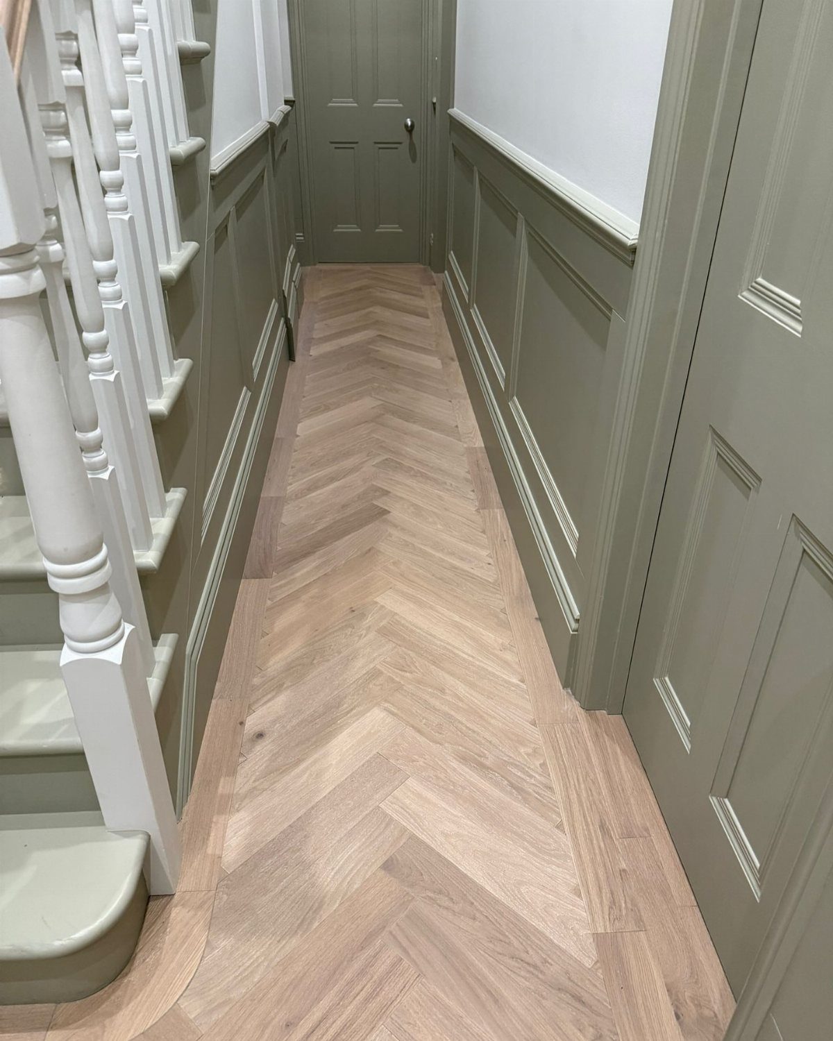 Light wood herringbone flooring in a narrow hallway with sage green wainscoting and white stairs.