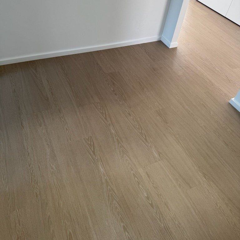Light brown wood plank flooring meeting a white wall and baseboard in a bright room.