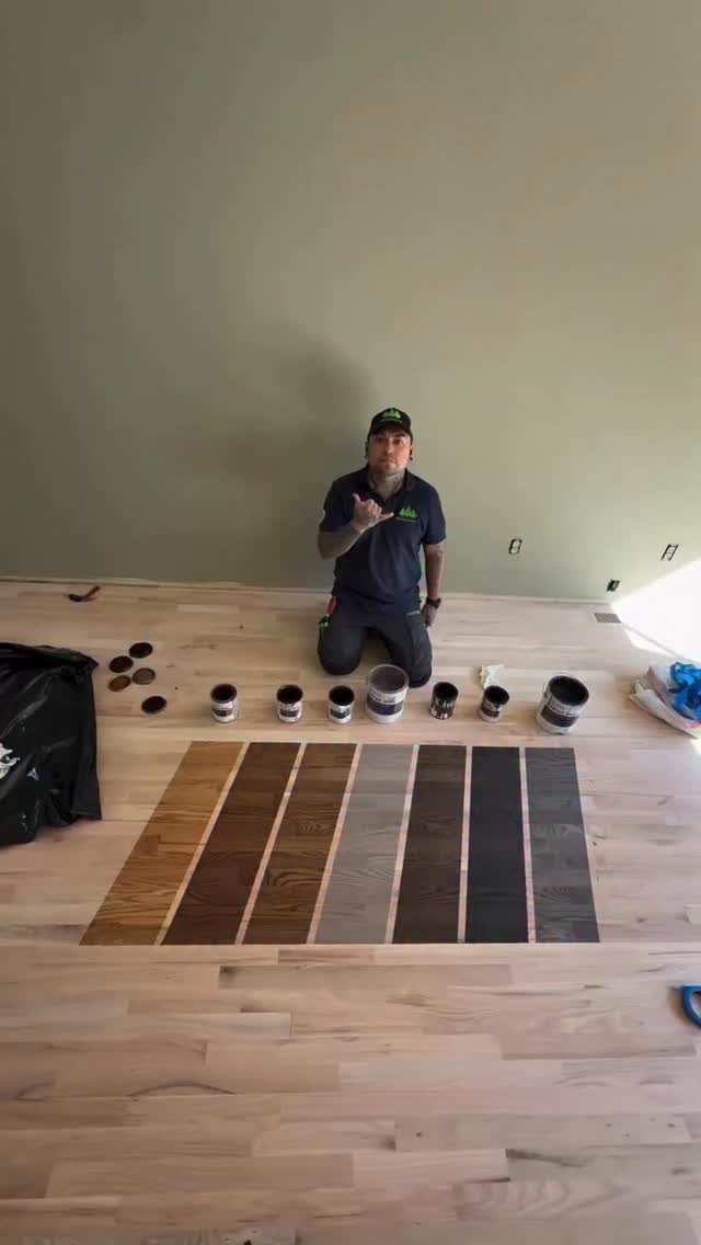 A man kneeling on a light wood floor next to various stain samples and paint cans.